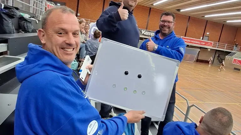 Drie CSV-begeleiders met een whiteboard op de tribune tijdens de Limburg Handball Cup.