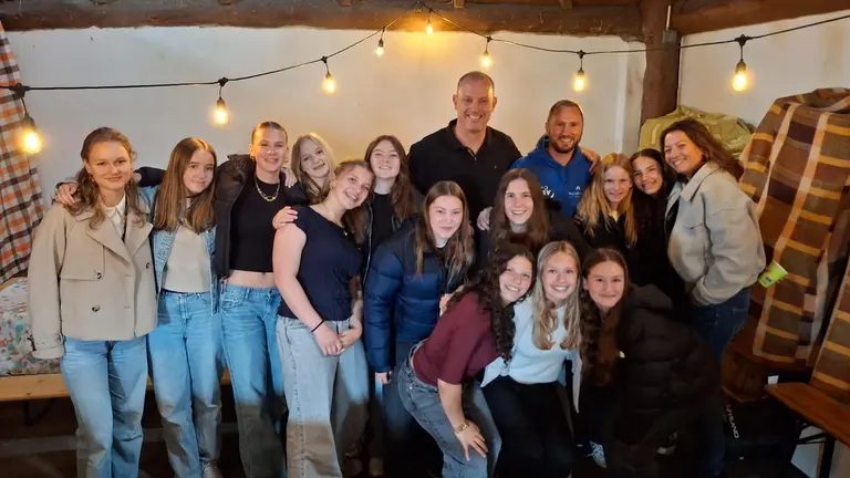 Groepsfoto van het team met ouders en begeleiders in een schuur met sfeerverlichting bij de eindejaarsafsluiting.