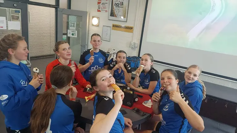 Speelsters van de MB2 eten ijsjes en snacks aan tafel in de kantine tijdens het toernooi.
